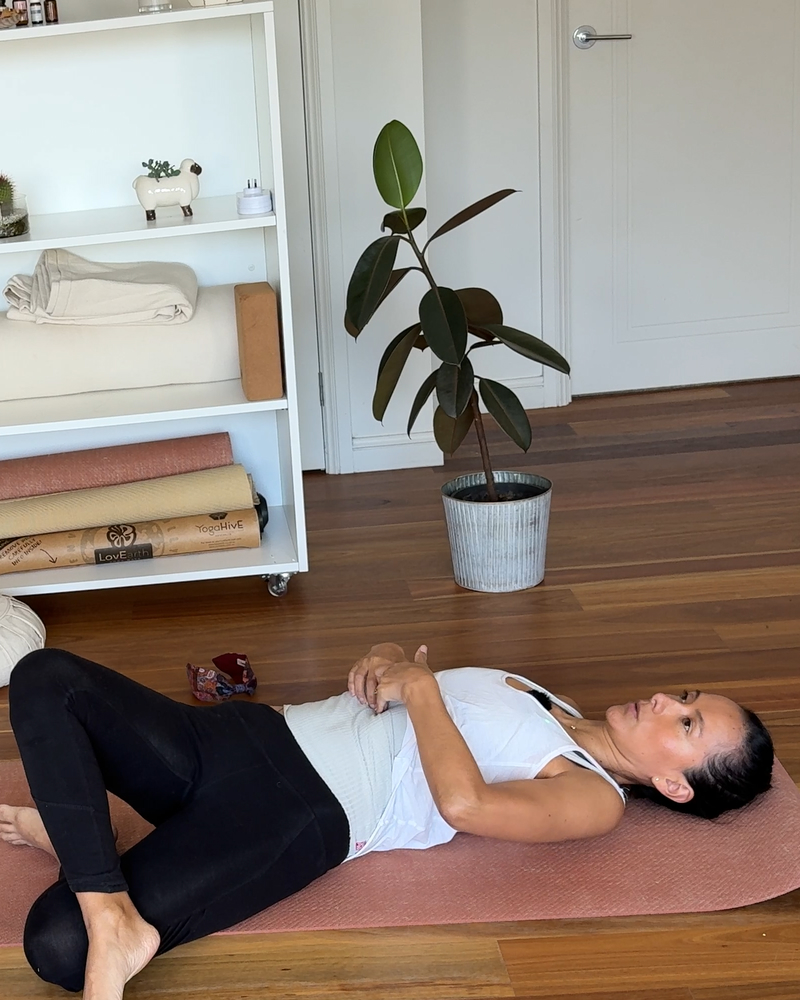 Person lying on a yoga mat in a bright practice space, resting with hands on their abdomen